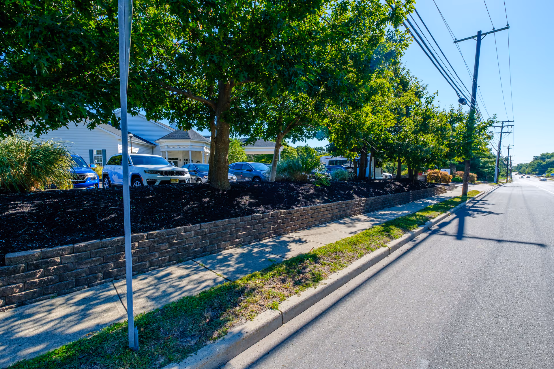 Street view showing a sidewalk with a low brick retaining wall and trees providing shade. Behind the wall, several parked cars and part of a white building with a porch are visible. Utility poles and power lines run along the road on the right side under a clear blue sky.