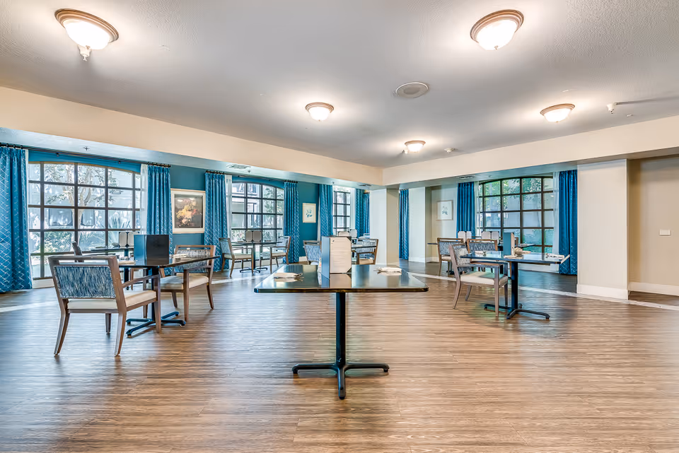 A spacious dining room in a senior living facility with wooden floors, multiple tables and chairs arranged neatly, large windows with blue curtains allowing natural light to fill the room, and soft ceiling lights providing additional illumination.