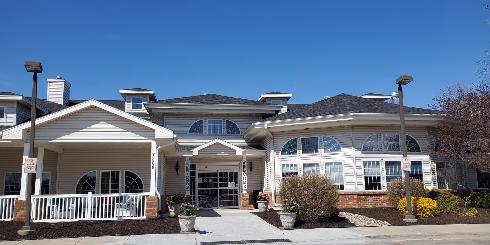 Front exterior view of Bailey Pointe Assisted Living at Van Dorn, showing a light-colored building with multiple windows, a covered entrance with a porch, potted plants, and clear blue sky above.