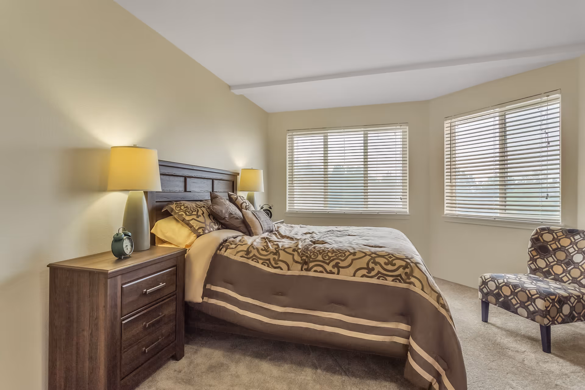 A cozy bedroom with a large bed featuring patterned bedding and several pillows. There are two bedside tables, each with a lamp, and an alarm clock on the left table. The room has beige walls, carpeted floor, and two large windows with blinds letting in natural light. A patterned armchair is placed near the windows.