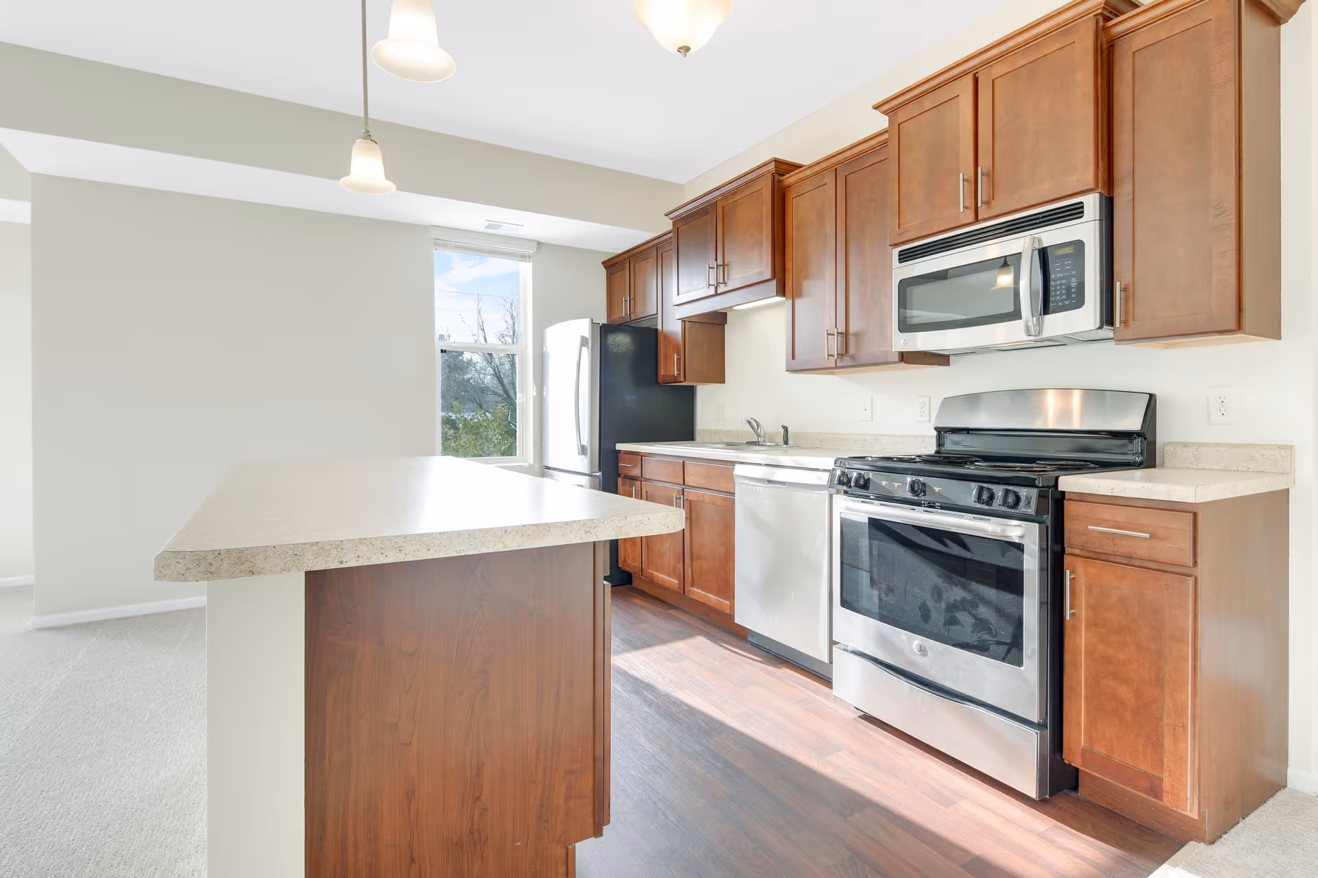 Bright kitchen with wooden cabinets, stainless steel appliances including a stove, microwave, dishwasher, and refrigerator. There is a large countertop island in the foreground and a window letting in natural light.