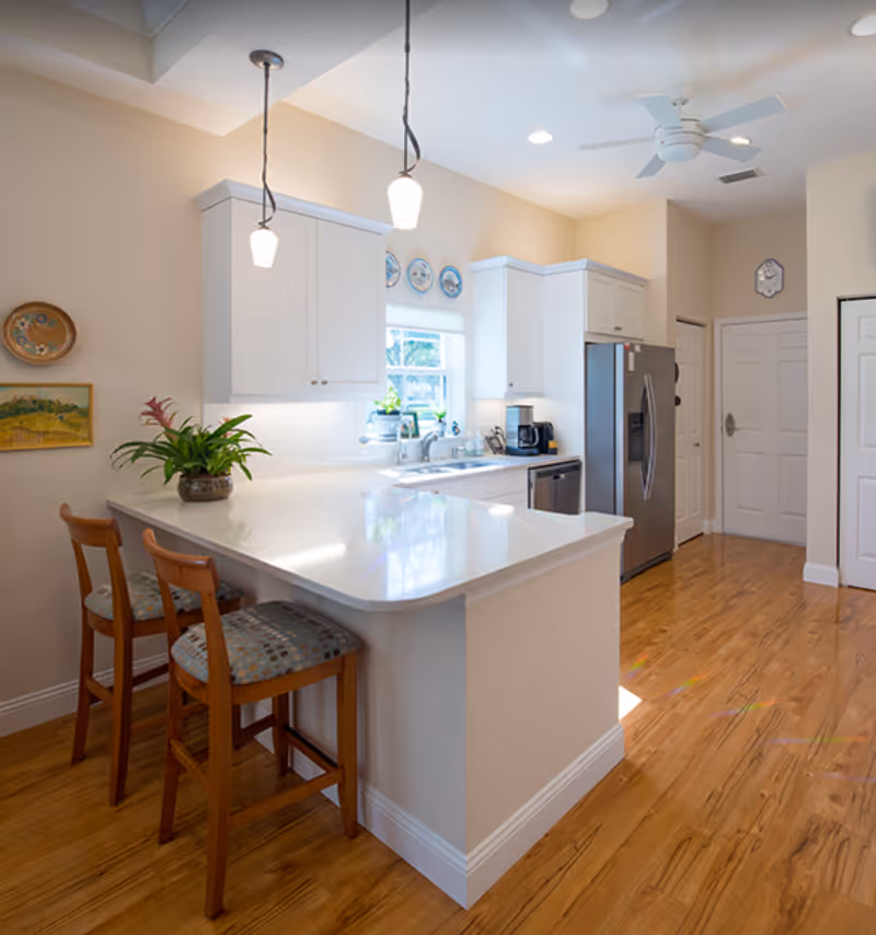 Bright and modern kitchen with white cabinets, a large white countertop island with two wooden chairs, pendant lights hanging above, a stainless steel refrigerator, a coffee maker, and a window above the sink letting in natural light. The floor is wooden and the walls are painted beige with some decorative plates and artwork.