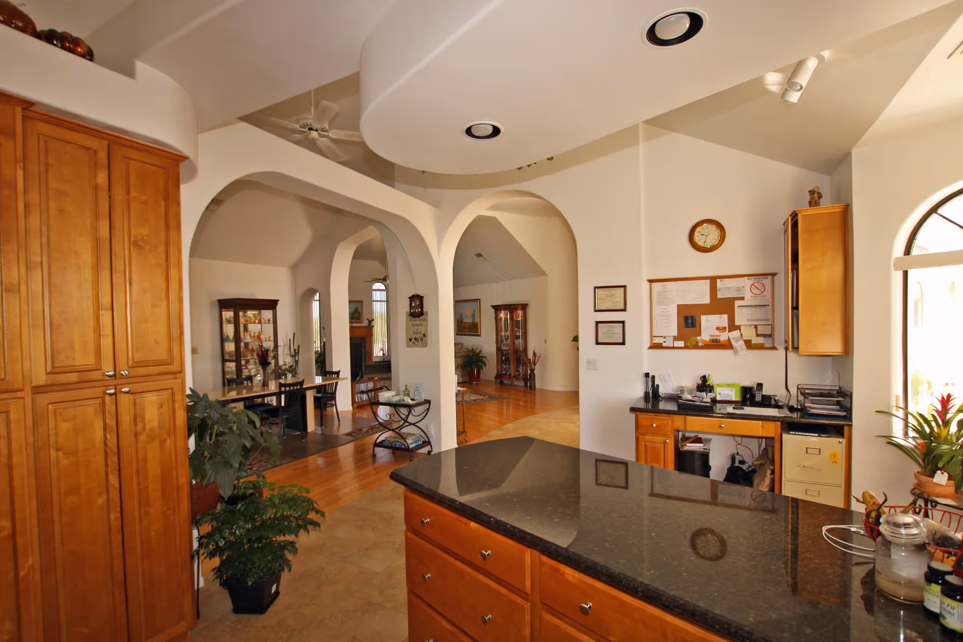 Interior view of a living space in Carefree Assisted Living, LLC showing a kitchen area with wooden cabinets and a black granite countertop island in the foreground. The background features arched doorways leading to a dining area with a table and chairs, a display cabinet, and a living room with a fireplace. The walls are white, and there are plants and decorative items throughout the space.