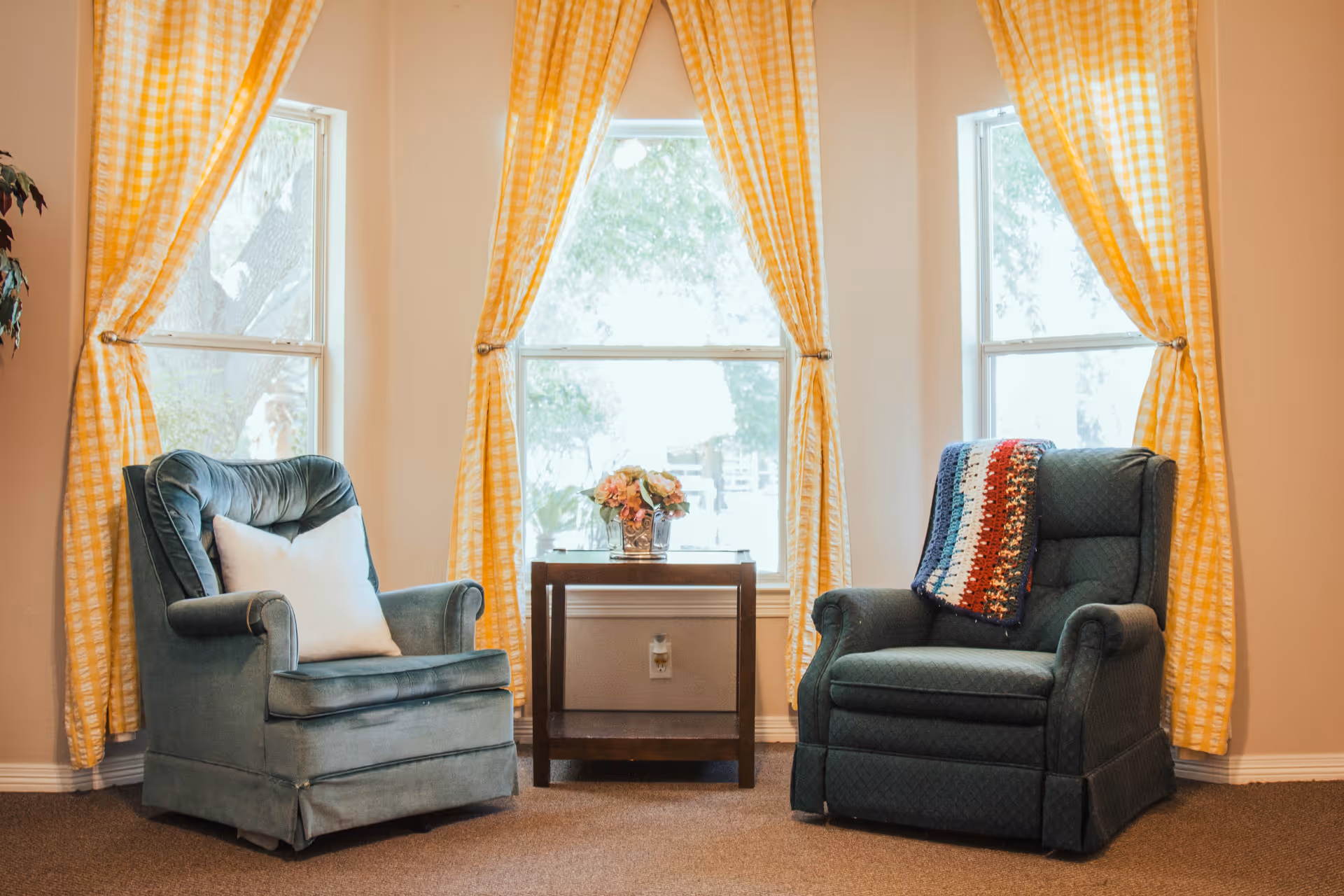 A cozy sitting area with two upholstered armchairs, one with a white pillow and the other with a colorful knitted throw, positioned on either side of a small wooden table with a vase of flowers. Behind them are three large windows with yellow checkered curtains tied back, allowing natural light to fill the room.