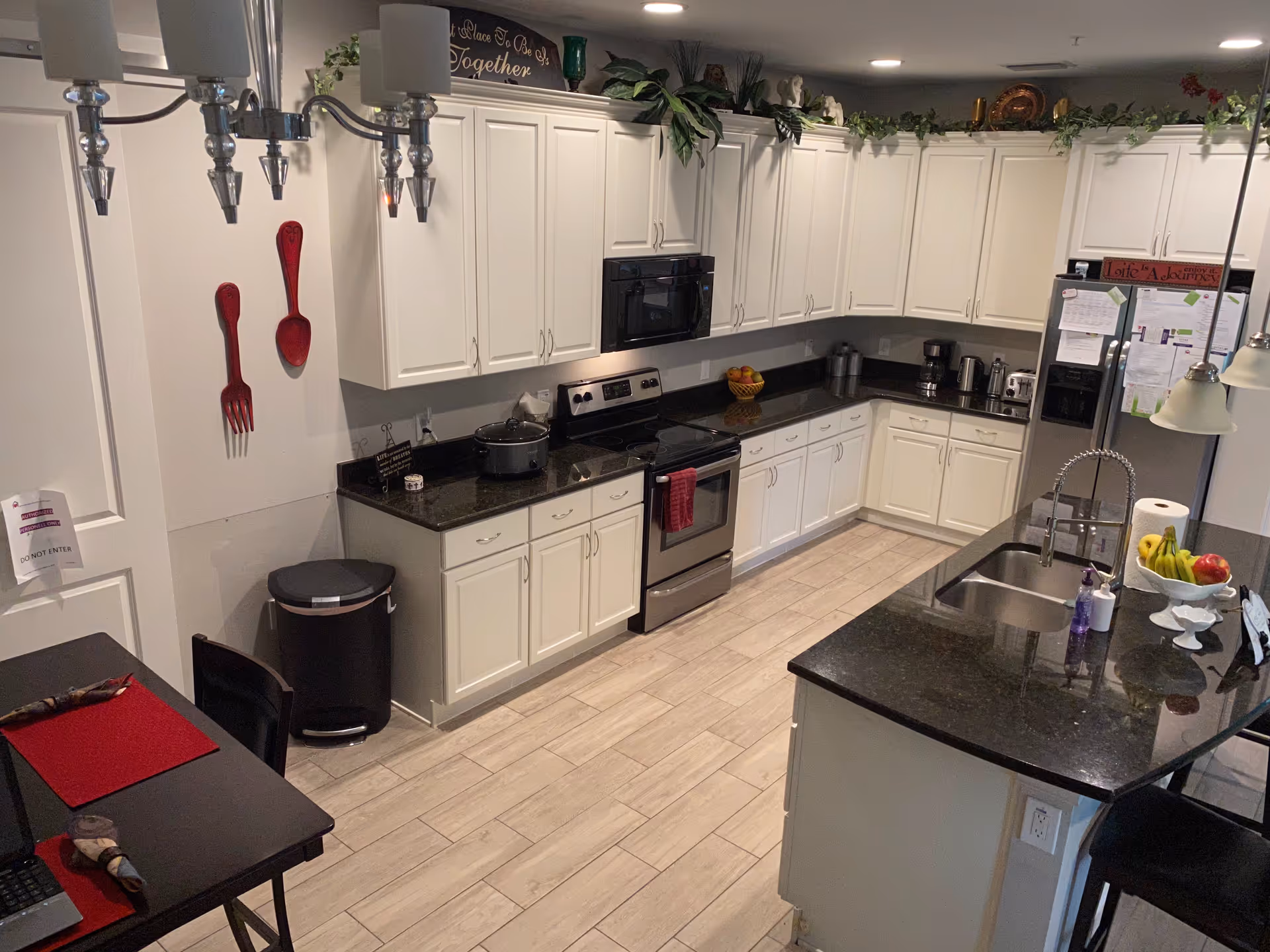 A modern kitchen with white cabinets, black granite countertops, and stainless steel appliances including a stove, microwave, and refrigerator. There is a kitchen island with a sink and a bowl of fruit, as well as a small dining table with red placemats and black chairs. Decorative plants and wall art are placed above the cabinets.