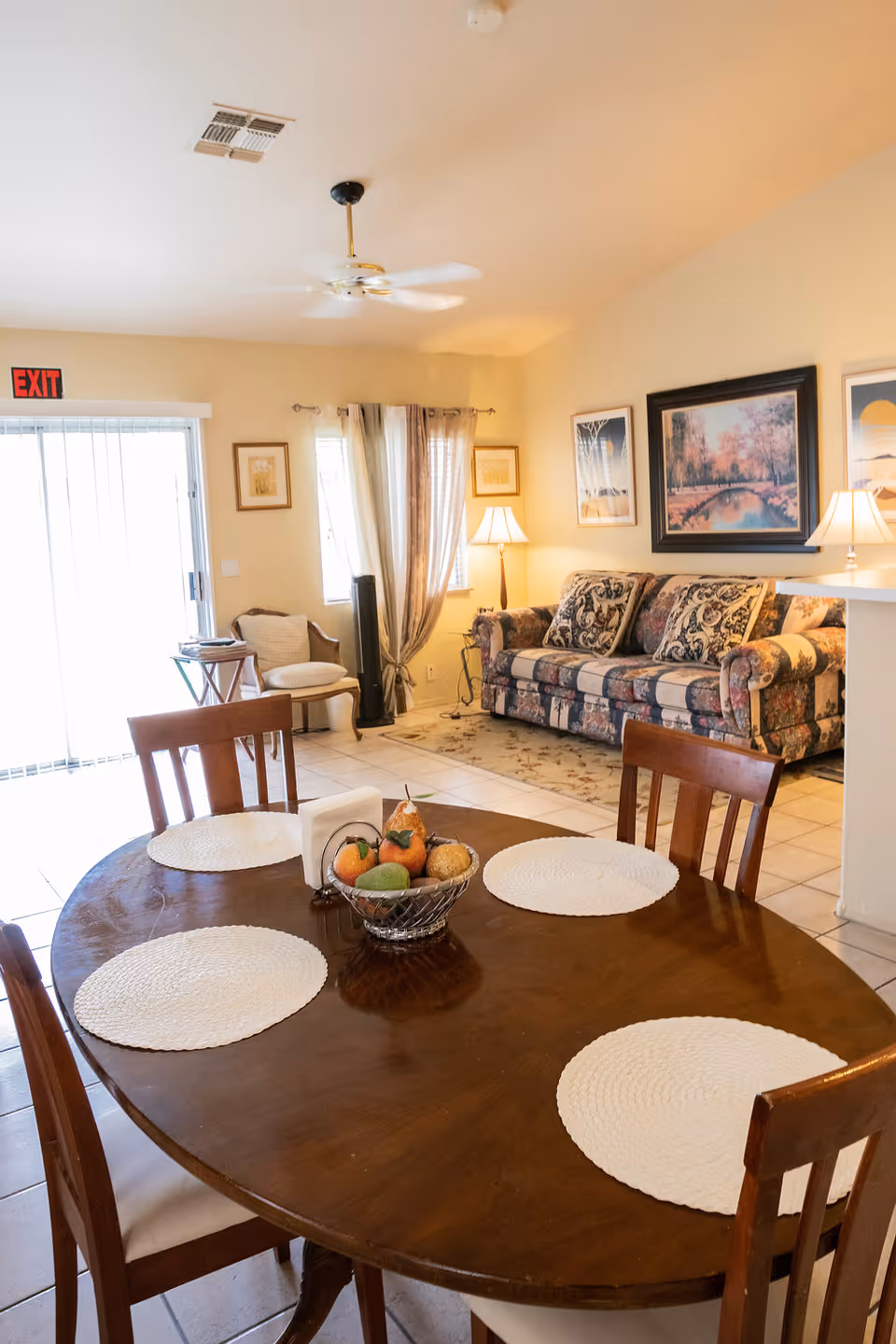 A cozy living and dining area with a wooden oval dining table set with four white placemats and a fruit basket centerpiece. In the background, there is a floral-patterned sofa with decorative pillows, a floor lamp, framed artwork on the walls, a small chair near a window with curtains, and a sliding glass door with an exit sign above it.