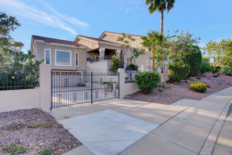 Exterior view of a residential-style building with beige stucco walls and a red tile roof, surrounded by a black wrought iron fence and gate. The landscape includes various green bushes, trees, and a palm tree, with a sidewalk and driveway in the foreground under a clear blue sky.