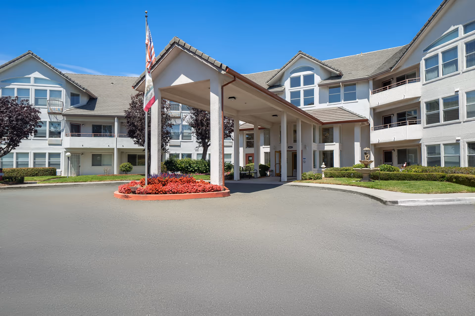 Front exterior view of a multi-story senior living facility with a covered entrance, landscaped flower beds, and American and California state flags on flagpoles under a clear blue sky.