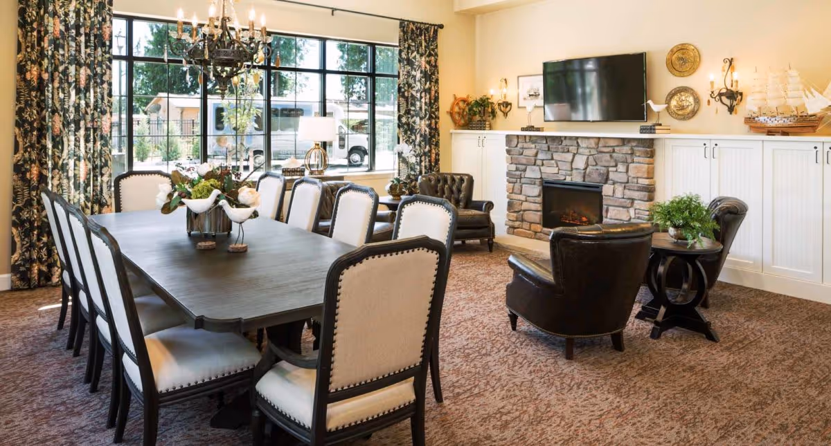 A cozy and elegant living room and dining area with a long dark wooden dining table surrounded by upholstered chairs. The room features a stone fireplace with a mounted flat-screen TV above it, two leather armchairs, decorative wall sconces, and a model ship on a cabinet. Large windows with floral curtains allow natural light to fill the space.