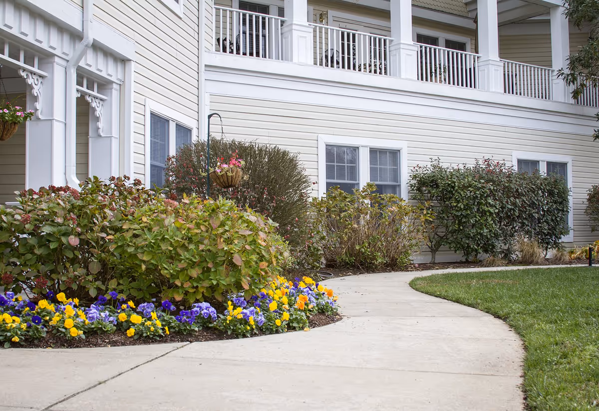 Curved concrete walkway bordered by flower beds and shrubs leading to the exterior of a light-colored multi-story residential building with a balcony.