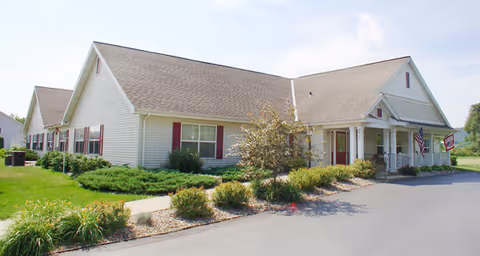 Exterior view of a single-story building with beige siding, a gable roof, and red shutters. The building is surrounded by green grass, shrubs, and small trees, with a paved driveway in front. An American flag is displayed near the entrance.