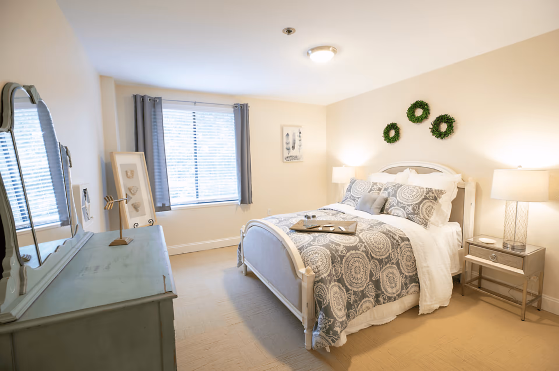 Sunny, tastefully decorated bedroom with a bed dressed in patterned bedding, nightstands with lamps, a dresser with mirror, and a window with curtains.