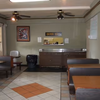 Interior view of a small dining area with tiled floor featuring a diamond pattern. There are wooden booths with dark cushions on the right and a table with chairs on the left. A counter with a sign above it that reads 'ORDER HERE' is visible at the back wall. Ceiling fans with lights are mounted on the ceiling, and a framed picture hangs on the left wall.