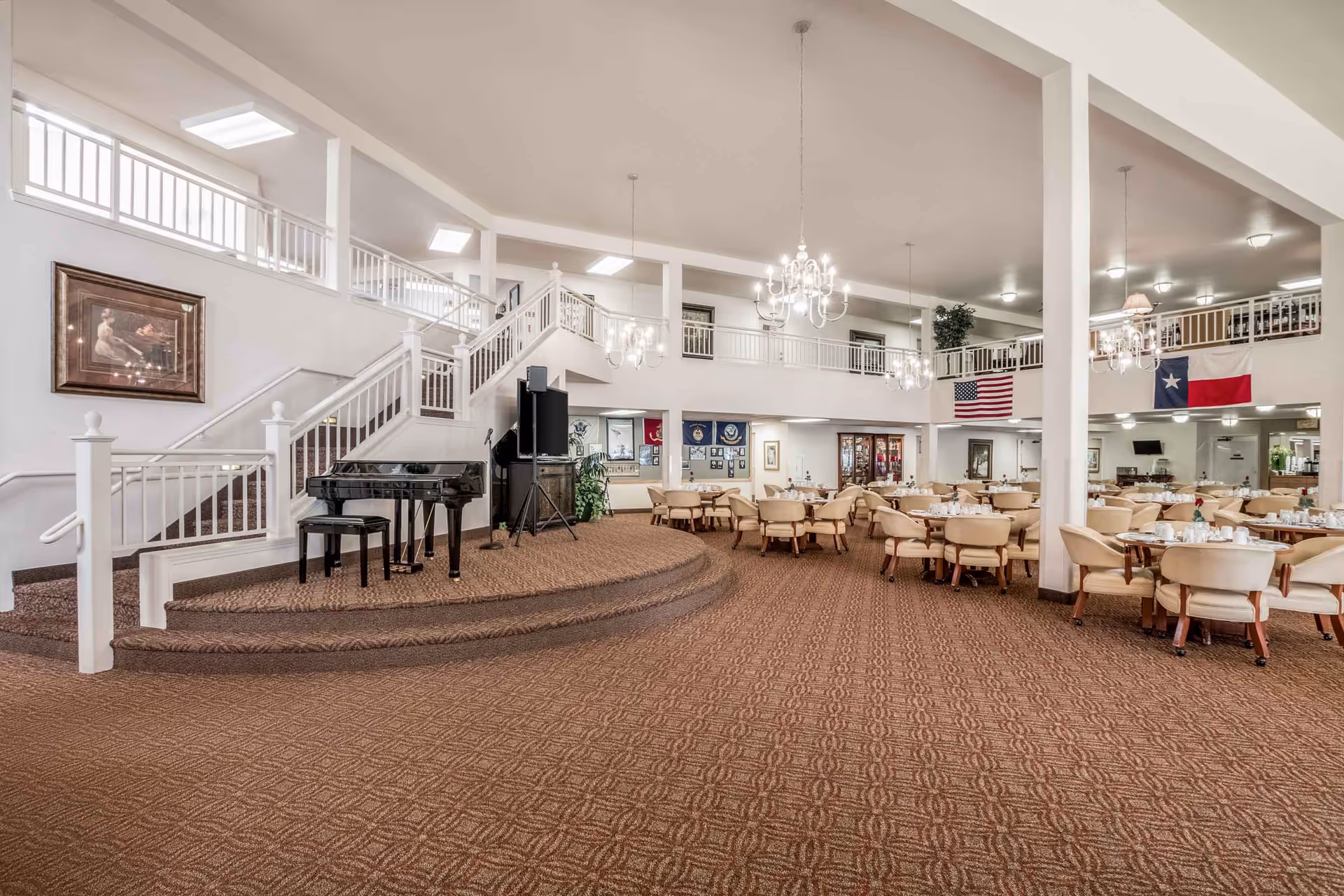 Spacious dining room with round tables and chairs, a small stage with a piano, and a staircase leading to an upper balcony.