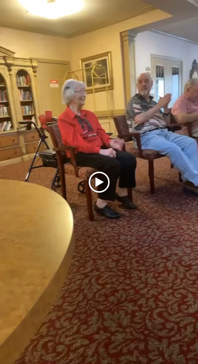 Three older adults seated in armchairs in a carpeted communal lounge with bookshelves and chairs.