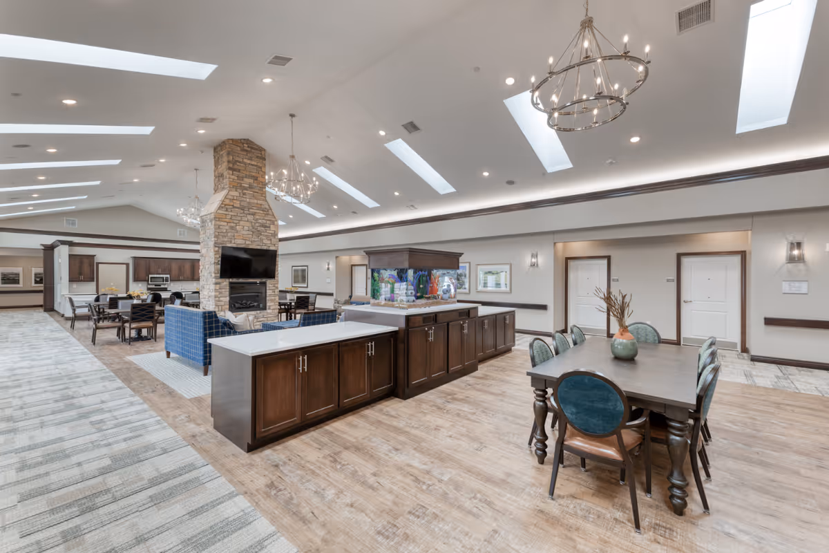 Spacious senior living common area featuring a large stone fireplace with a mounted TV, multiple seating arrangements including blue armchairs and dining tables, a large fish tank on a wooden cabinet, and skylights in the ceiling providing natural light.