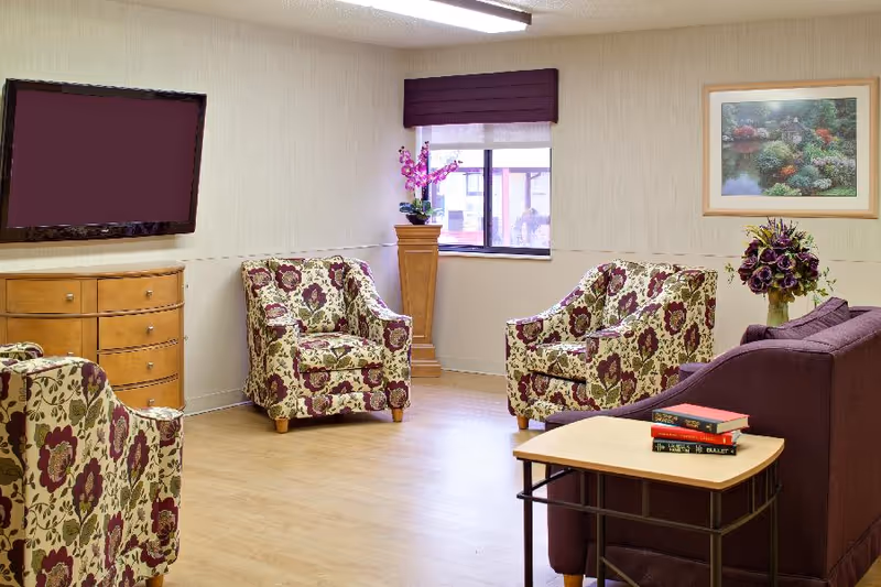 A cozy living room area with three floral-patterned armchairs and a purple sofa. A wooden dresser with a flat-screen TV mounted above it is on the left wall. A small wooden table with three books sits in front of the sofa. There is a window with a purple valance, a tall wooden plant stand with a purple orchid, and a framed picture of a garden scene on the wall.