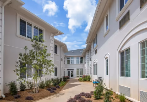 Outdoor walkway between two white buildings with multiple windows, small landscaped plants and bushes along the path, a bench with blue cushions on the right side, under a partly cloudy blue sky.