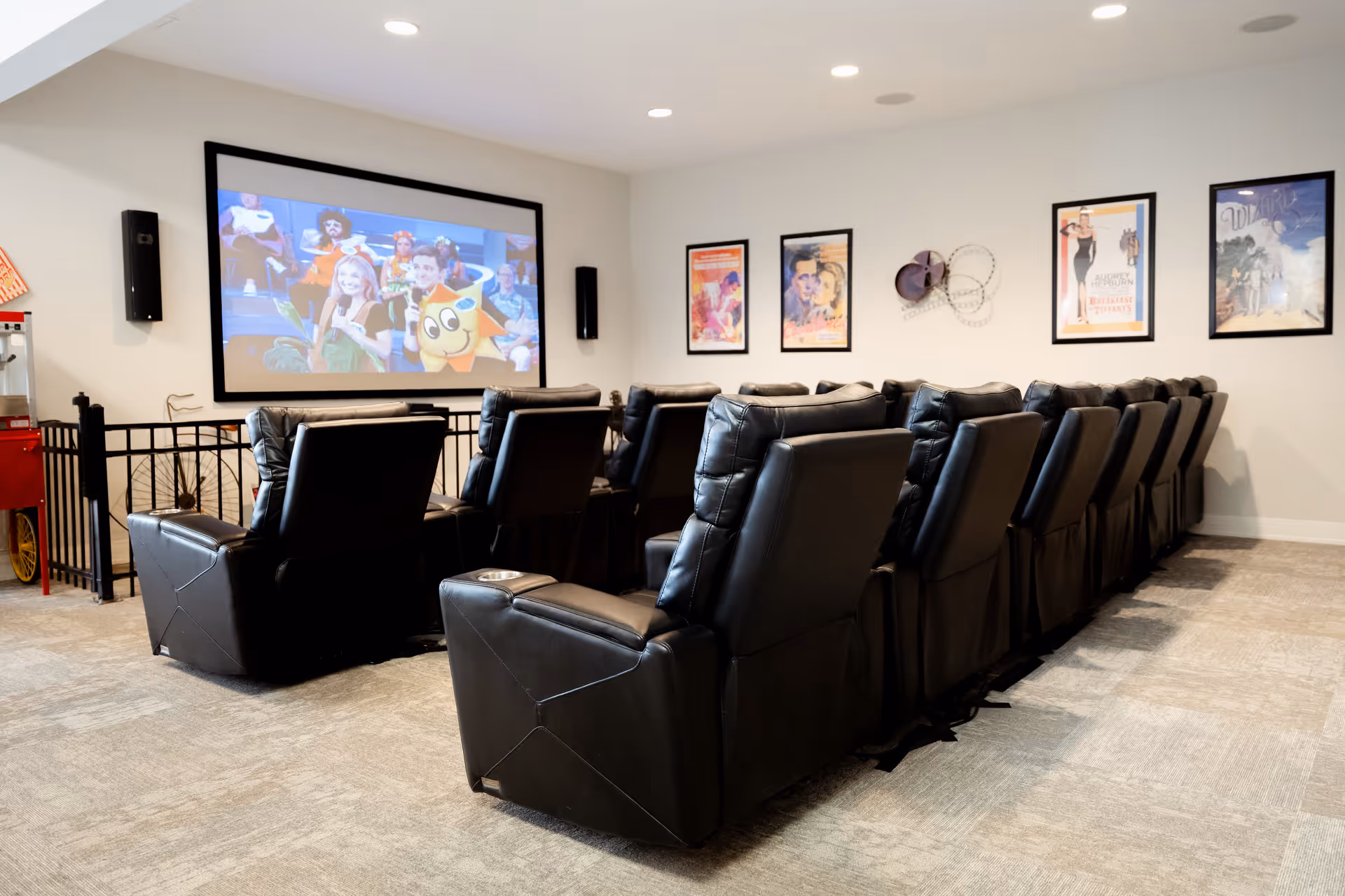A cozy movie theater room with two rows of black leather recliner chairs facing a large screen showing a colorful cartoon. The walls are decorated with framed movie posters and a popcorn machine is visible in the corner.