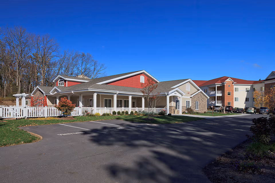 Exterior view of a senior living facility named Crestwood on a clear sunny day. The building features a combination of beige, red, and stone facade with a white picket fence and a covered porch. There are several parked cars and trees with autumn foliage surrounding the property.