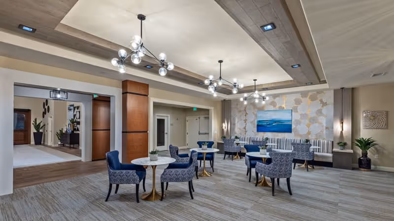 A spacious and modern common area with multiple small round tables and blue patterned chairs arranged around them. The room features a high ceiling with recessed lighting and contemporary chandeliers. There is a decorative wall with a large blue and white painting, and plants are placed around the room for a welcoming atmosphere.
