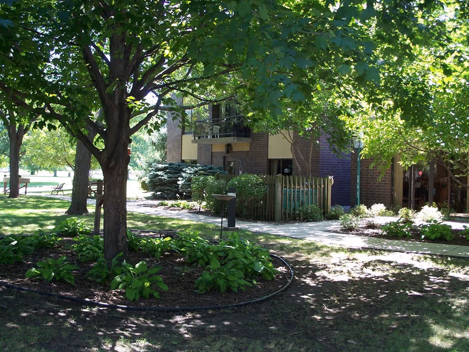 A shaded outdoor garden area with large trees, green plants, and a pathway leading to a brick building with balconies and windows. There are benches in the background and a small fenced area near the building.