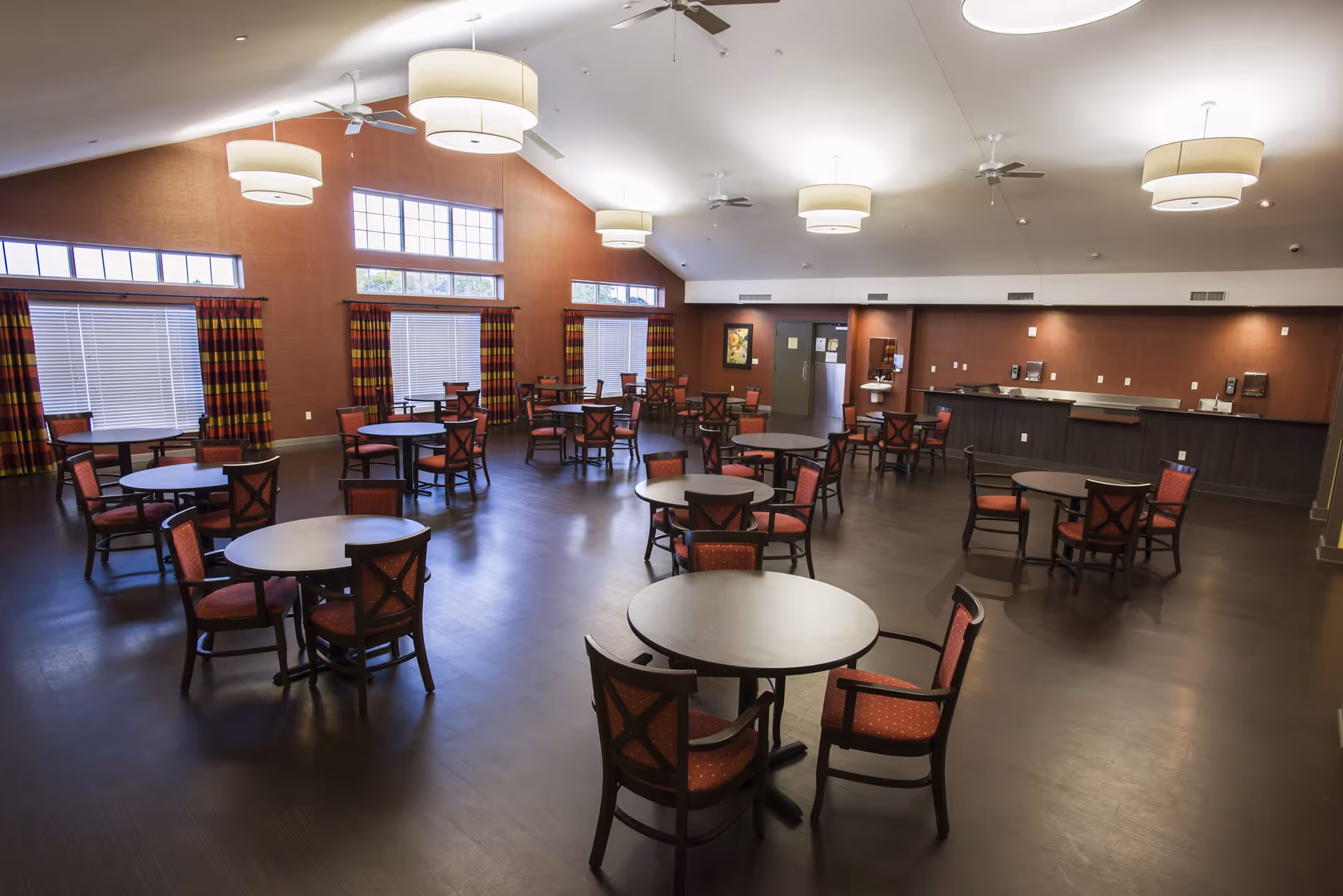A spacious dining room with multiple round tables and red cushioned chairs arranged neatly. The room has large windows with striped curtains, ceiling fans, and modern hanging light fixtures. The walls are painted a warm brown color, and there is a counter area along one wall with sinks and dispensers.