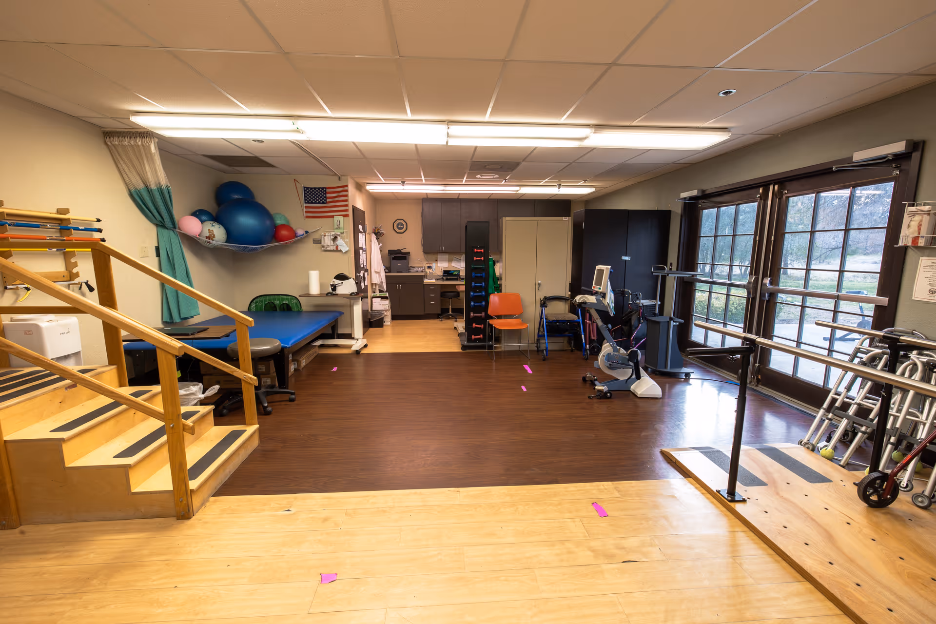A physical therapy room in a senior living facility with wooden stairs for rehabilitation, exercise balls on a shelf, a treatment table, various mobility aids including walkers and wheelchairs, parallel bars for walking practice, and large windows letting in natural light.