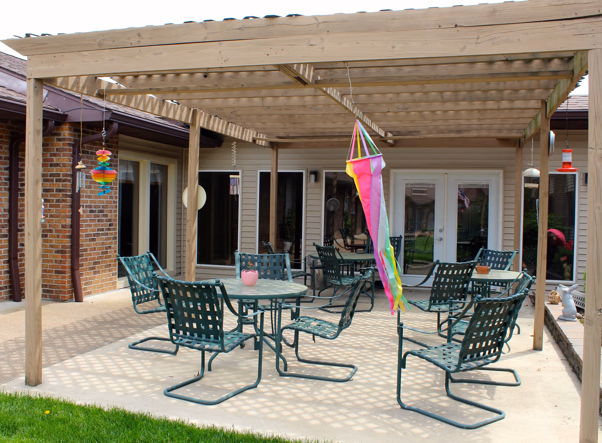 Outdoor patio area with a wooden pergola overhead, featuring several green metal chairs and round tables. Colorful wind chimes and a bright multicolored windsock hang from the pergola. The patio is adjacent to a building with brick and siding walls and glass doors and windows.