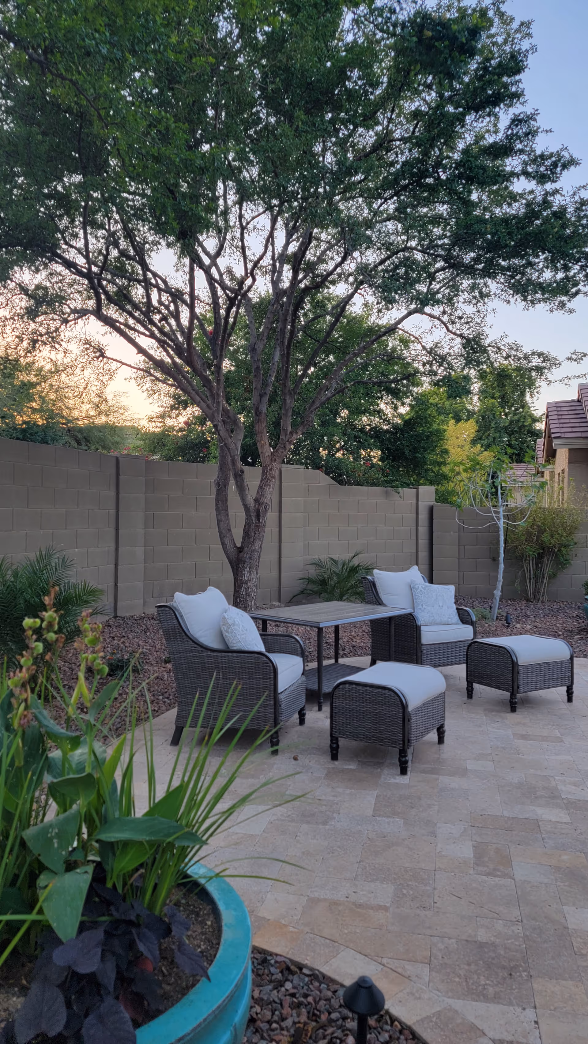 Outdoor patio area with two cushioned wicker chairs, two matching ottomans, and a small table set on a tiled floor. The patio is surrounded by a low stone wall and landscaping with trees and plants, under a clear sky at sunset.
