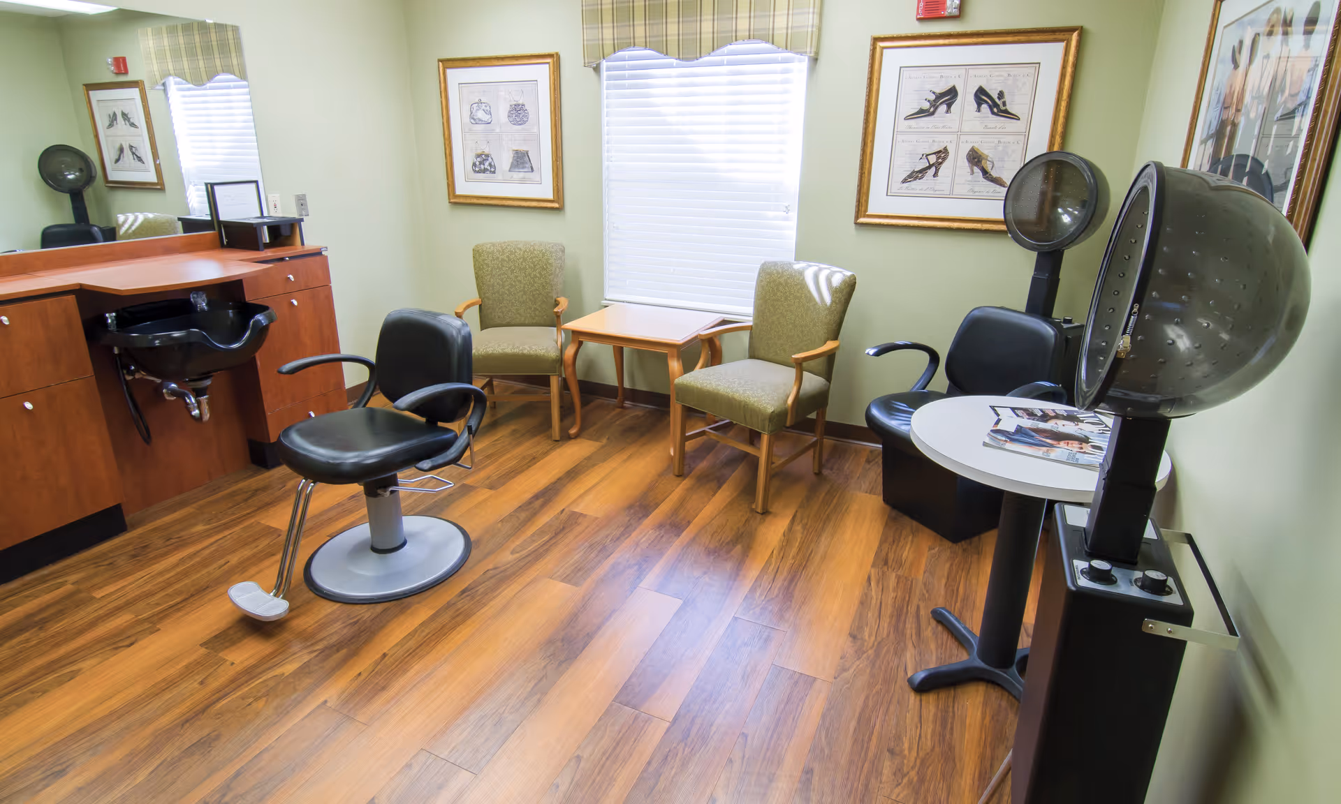 Interior view of a salon room with wooden flooring, two black salon chairs, a black hair washing sink, two green armchairs with wooden arms, a small wooden table between the armchairs, a round white table with magazines, and two hair dryers. The walls are light green with framed artwork and a window with blinds.