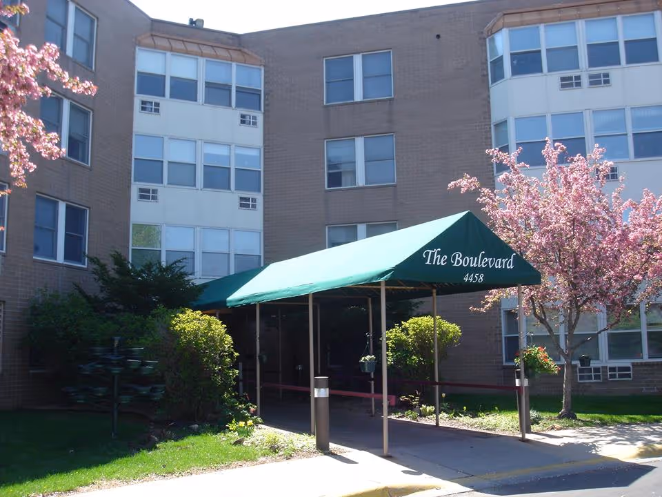 Exterior view of a multi-story brick building with a green canopy entrance labeled 'The Boulevard 4458'. There are flowering trees and shrubs around the entrance, and the building has multiple windows.