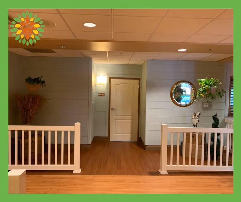 Interior hallway area with wooden flooring, light blue paneled walls, a white door in the center, white railings on both sides, a round mirror on the right wall, a hanging plant, and decorative items including a potted plant and a rooster figurine.
