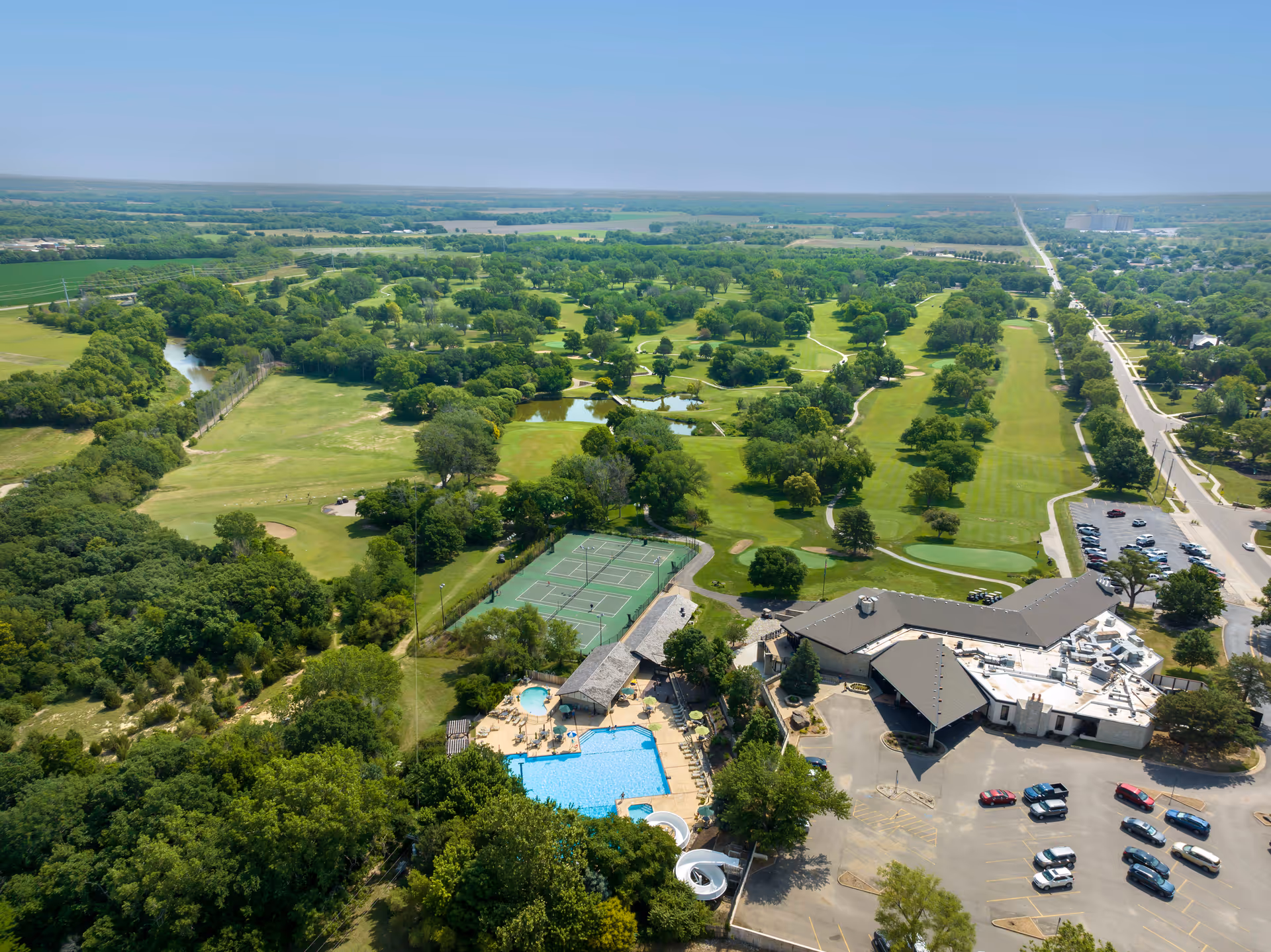 Aerial view of a senior living facility named Brookdale Salina Fairdale showing a large building with a parking lot, an outdoor swimming pool, tennis courts, and surrounding green spaces including a golf course with trees and walking paths.