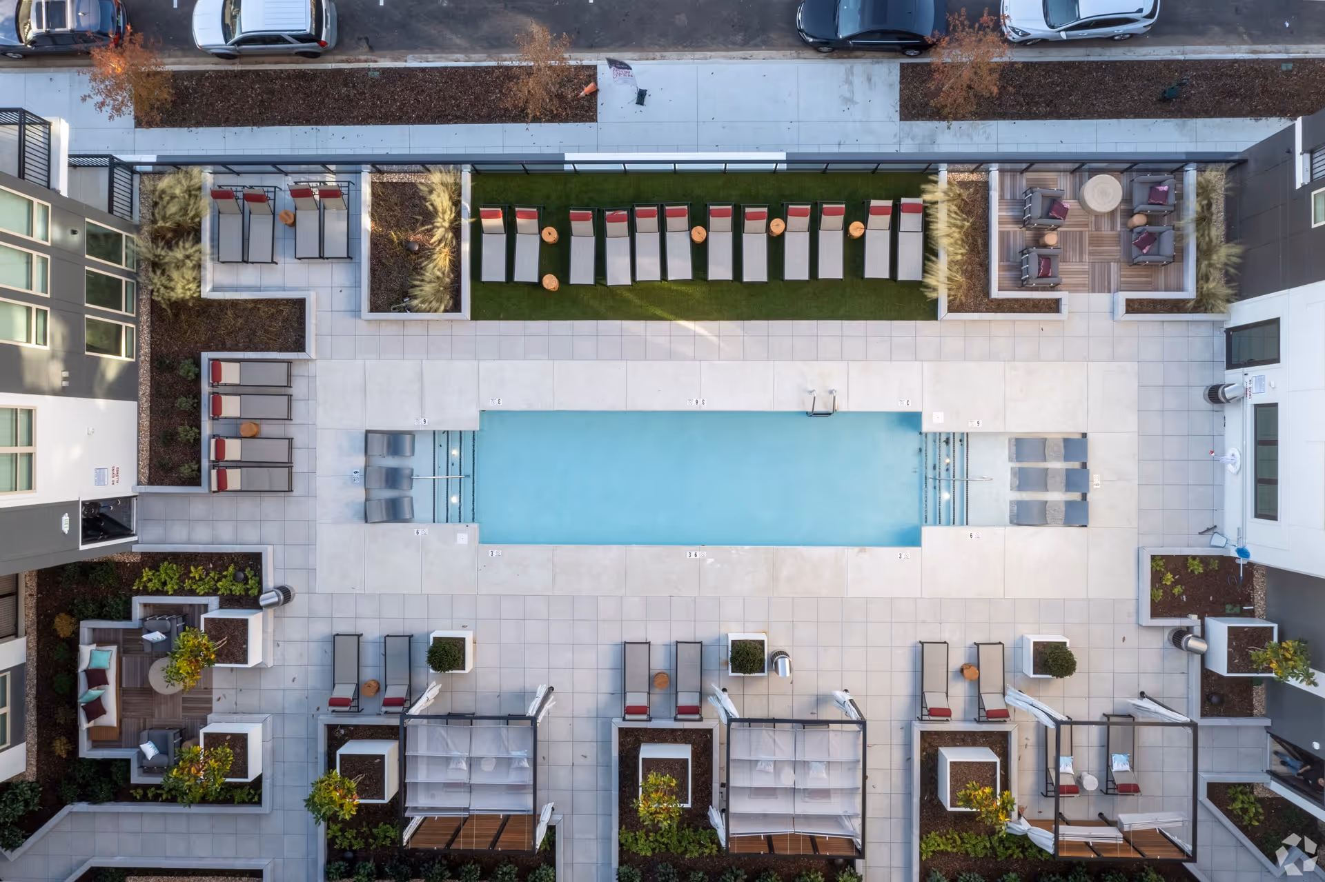 Aerial view of an outdoor swimming pool area at Upside Birmingham, surrounded by lounge chairs with red cushions, small wooden tables, cabanas, and seating areas with chairs and cushions. The pool is rectangular with steps on both ends, and the surrounding area is paved with light-colored tiles. There are landscaped garden beds with plants and trees around the pool area, and parked cars are visible on the street beyond the fenced perimeter.