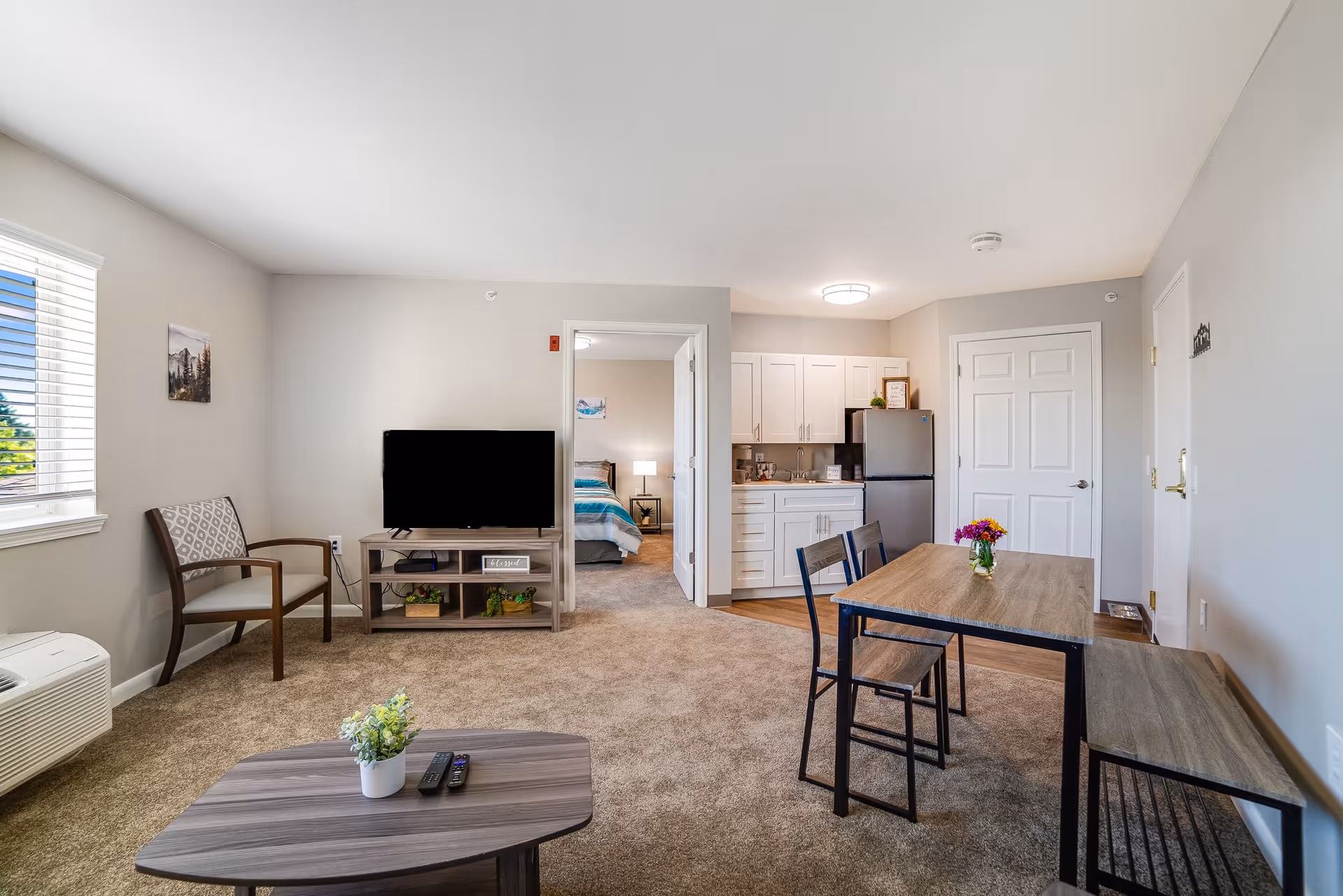 A bright and clean living space in a senior living facility featuring a small dining table with two chairs and a bench, a coffee table with a small plant and remote controls, a TV on a stand, a single armchair, and a kitchenette with white cabinets and a refrigerator. A bedroom with a bed and nightstand is visible through an open doorway.