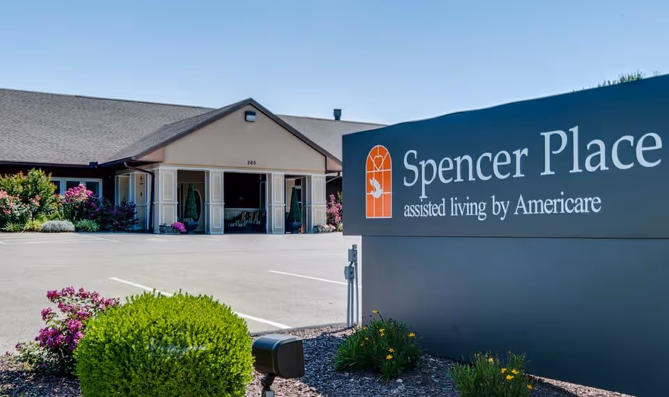 Exterior view of Spencer Place assisted living facility with a large sign in the foreground displaying the facility name and logo. The building has a beige facade with a peaked roof, surrounded by landscaped bushes and flowers under a clear blue sky.