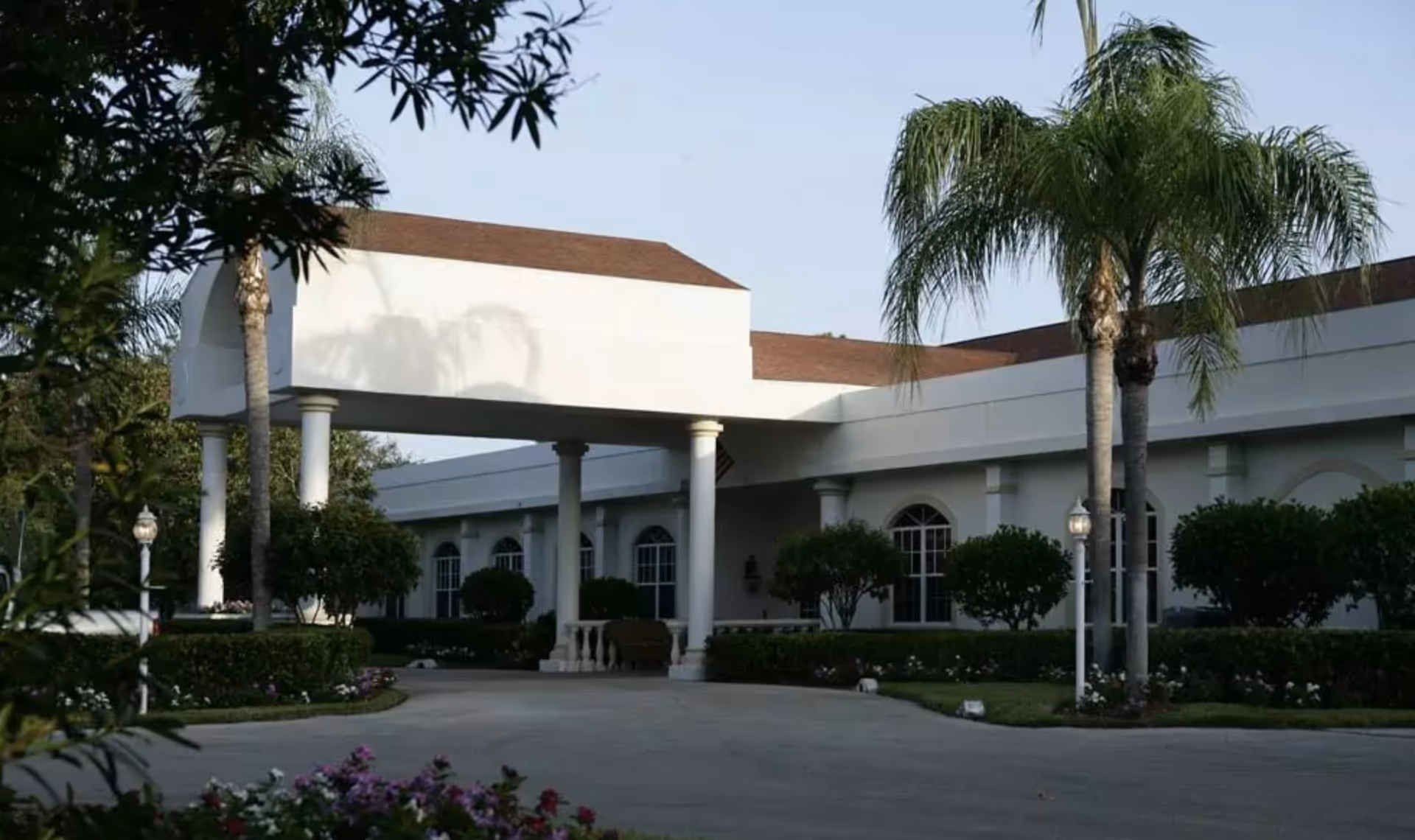 Exterior view of a white building with a covered entrance supported by columns, surrounded by palm trees, bushes, and flowers under a clear sky.