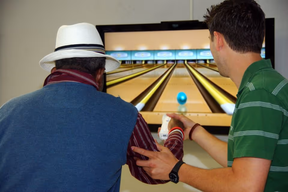 Two men playing a virtual bowling game on a large screen. One man, wearing a white hat and blue vest, is holding a game controller with assistance from the other man in a green striped shirt.