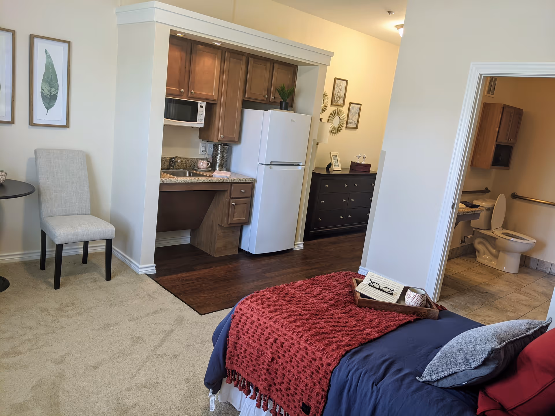 Interior view of a senior living facility room showing a bed with a red blanket and pillows, a small kitchenette with a refrigerator, microwave, and sink, a chair next to a small round table, and a bathroom with a toilet and grab bars visible through an open door.