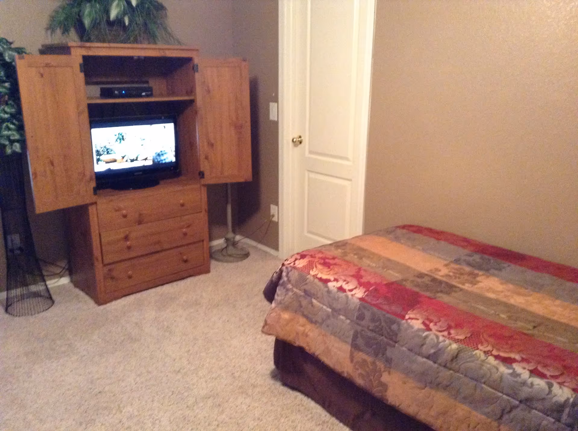 A bedroom with a bed covered in a multicolored quilt with floral patterns, a wooden armoire with open doors revealing a television inside, a beige carpeted floor, a closed white door, and a potted plant in the corner.