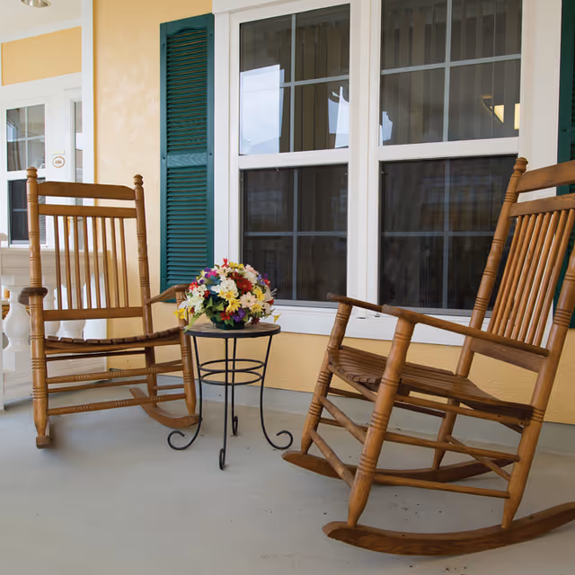 Two wooden rocking chairs on a covered porch with a small round table holding a bouquet in front of windows.