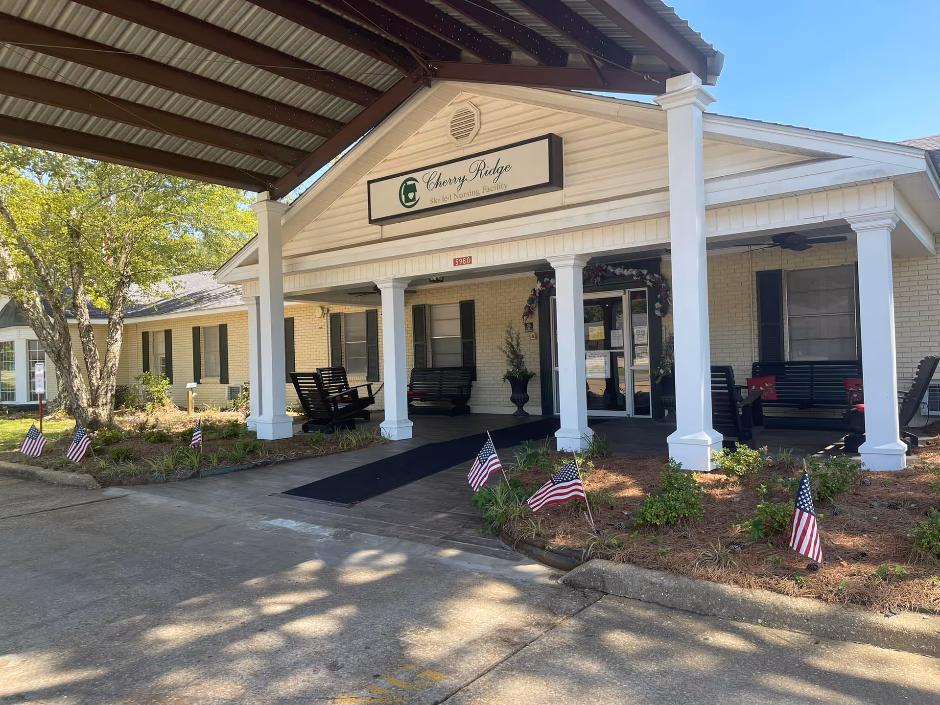 Front entrance of Cherry Ridge Skilled Nursing Facility with a covered porch supported by white columns. There are black benches and rocking chairs on the porch, small American flags planted in the garden beds, and a tree to the left. The building has light-colored brick walls and dark shutters on the windows.