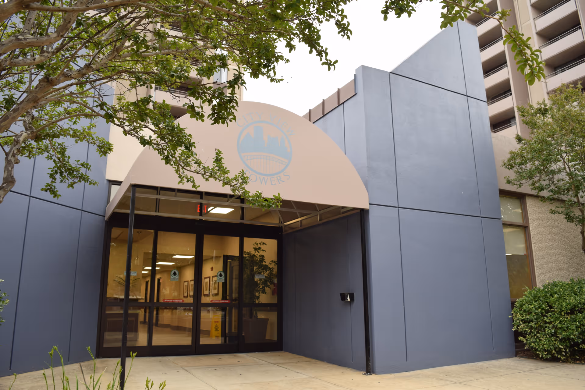 Entrance to City View Towers Senior Apartments featuring glass double doors under a beige awning with the facility's logo. The building exterior is gray with some greenery and trees around the entrance.