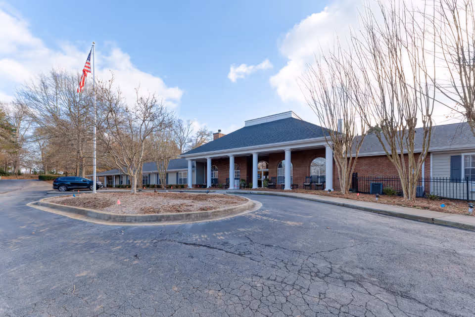 Front entrance of a single-story brick senior living facility with columns, a circular driveway, and an American flag on a flagpole.