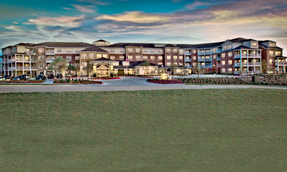 Front exterior view of a large multi-story retirement facility with balconies, a central entrance and a wide lawn in the foreground.