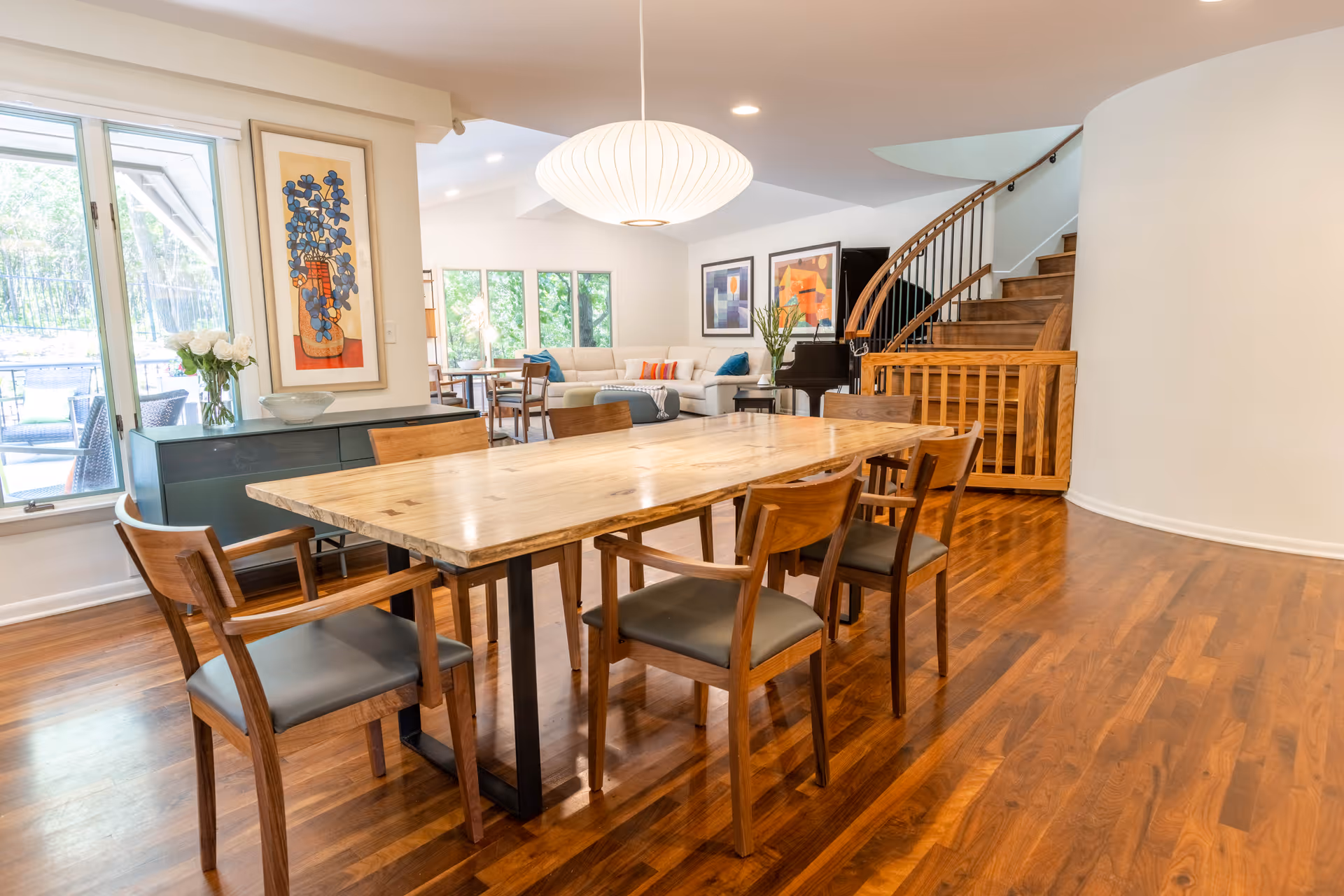Spacious open dining area with a large wooden table and chairs, hardwood floors, a modern pendant light, and a view into a living room and staircase.