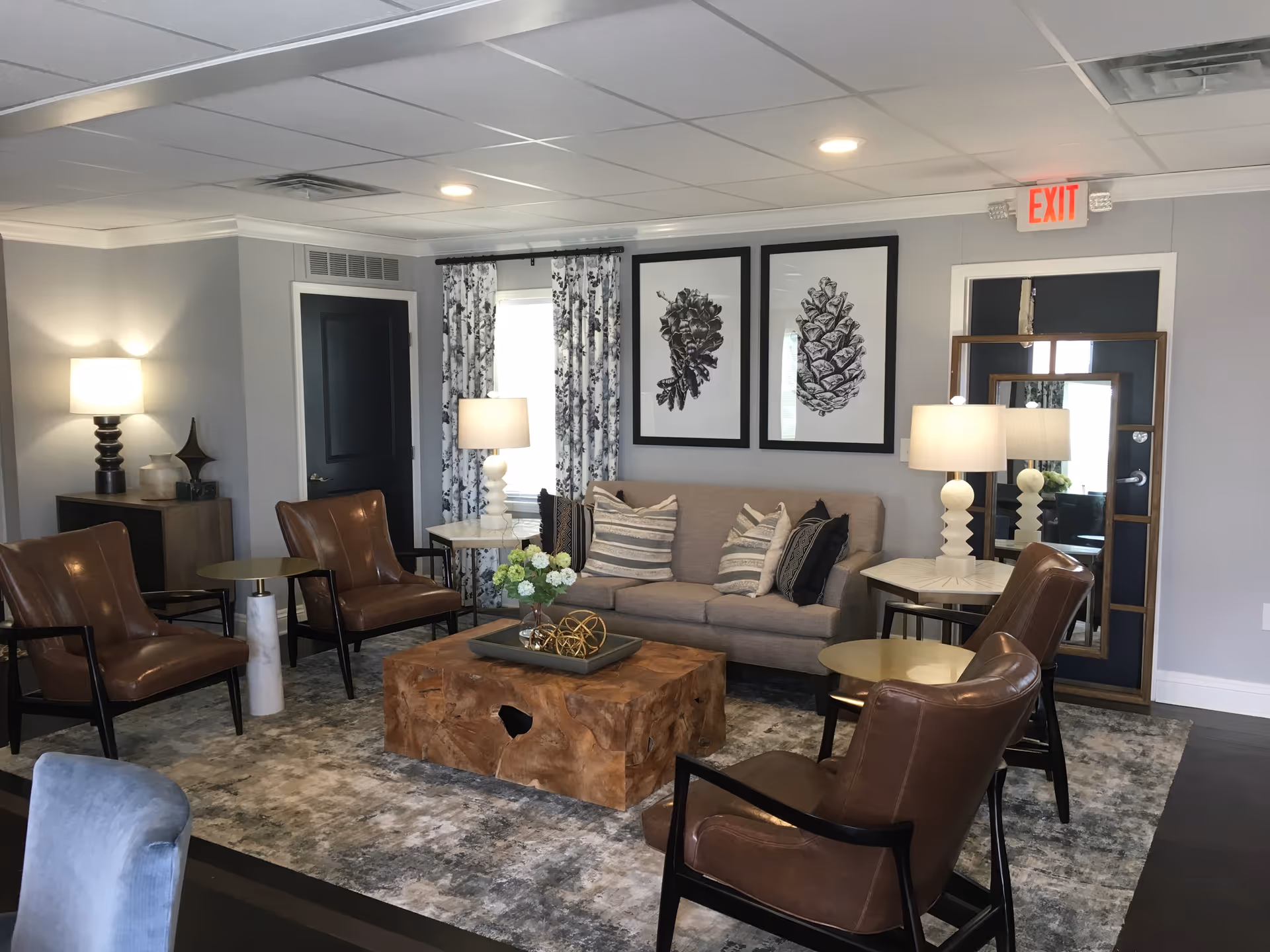 A cozy senior living facility lounge area with a beige sofa adorned with striped and patterned pillows, surrounded by four brown leather armchairs. The room features two side tables with white lamps, a wooden coffee table with decorative items, a large area rug, and two framed pinecone artworks on the wall. There is a window with patterned curtains and a door with a mirror leaning against it. The ceiling has recessed lighting and an exit sign above the door.