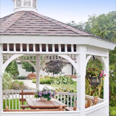 White wooden gazebo with a picnic table, potted flowers, and surrounding landscaped garden.