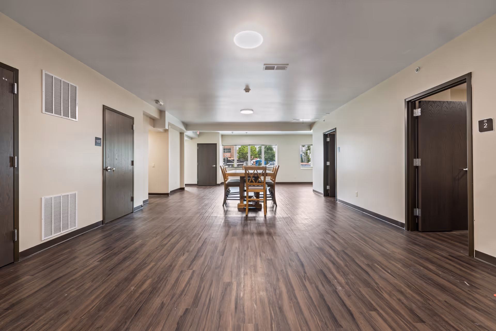 A spacious interior room with wood flooring and beige walls, featuring a wooden table with chairs in the center near a window. Several dark wooden doors are visible along the walls, and the ceiling has round light fixtures.